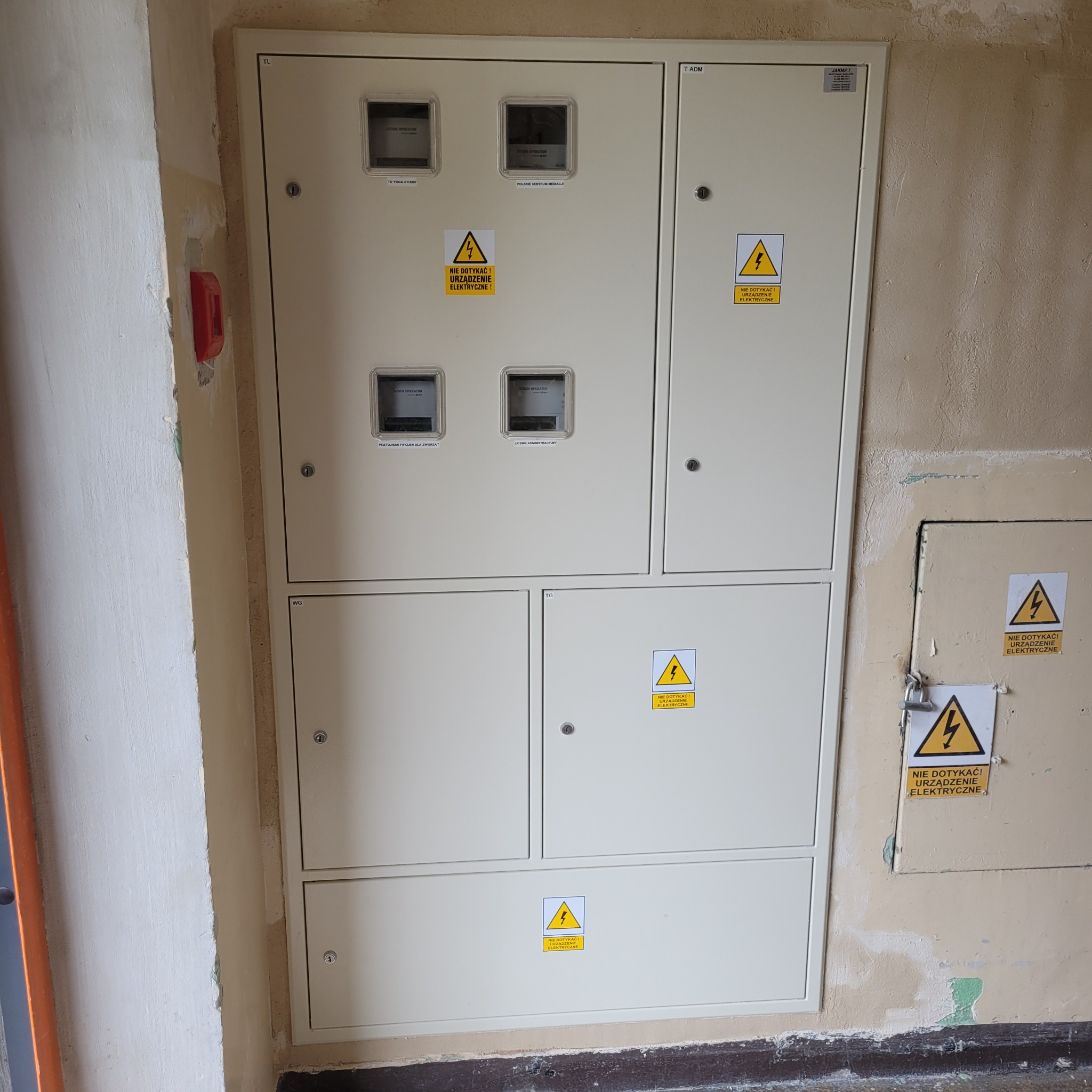 Rozdzielnica elektryczna z licznikami w budynku, z żółtymi naklejkami ostrzegawczymi. Widoczne ślady użytkowania na ścianie.