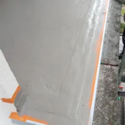 Szara, świeżo nałożona gładź na balkonie, zabezpieczona taśmą malarską, widok z góry, z fragmentem rusztowania i osoby w rękawicach w prawym górnym rogu.