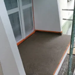 Balkon w trakcie prac: świeżo wylana wylewka, zabezpieczone okno folią, pomalowane ściany. Widoczne rusztowanie i taśma zabezpieczająca.