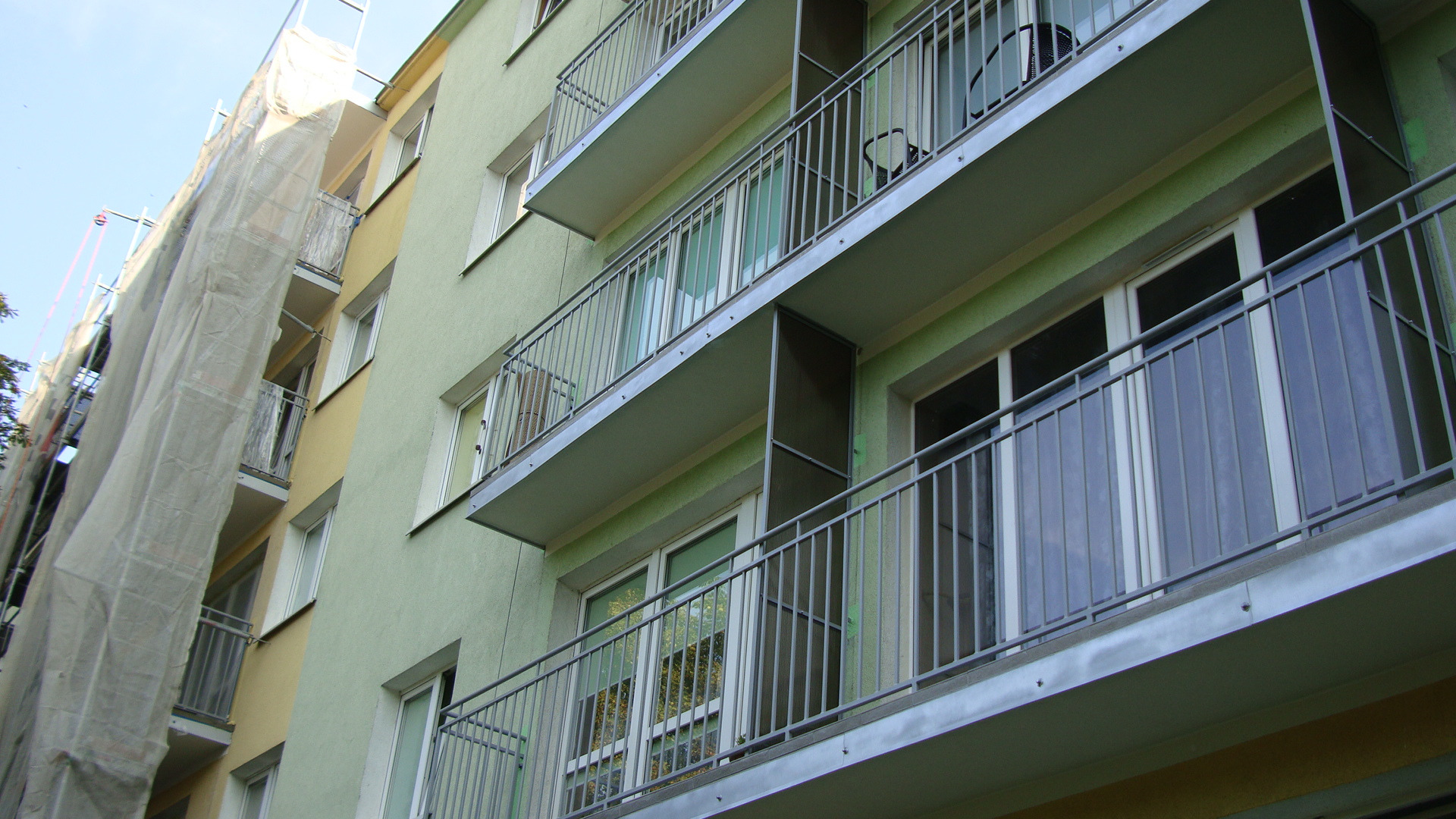 Budynek wielorodzinny z balkonami i fragmentem elewacji w trakcie prac remontowych, osłoniętej siatką ochronną. Widok z dołu, na pierwszym planie balkony.