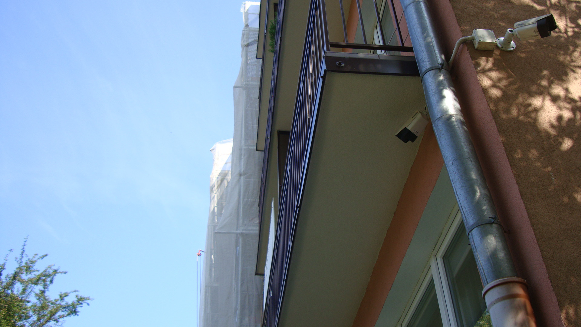 Budynek w trakcie renowacji elewacji, rusztowania osłonięte siatką, widoczne balkony i kamery monitoringu. Ujęcie z dołu, błękitne niebo w tle.