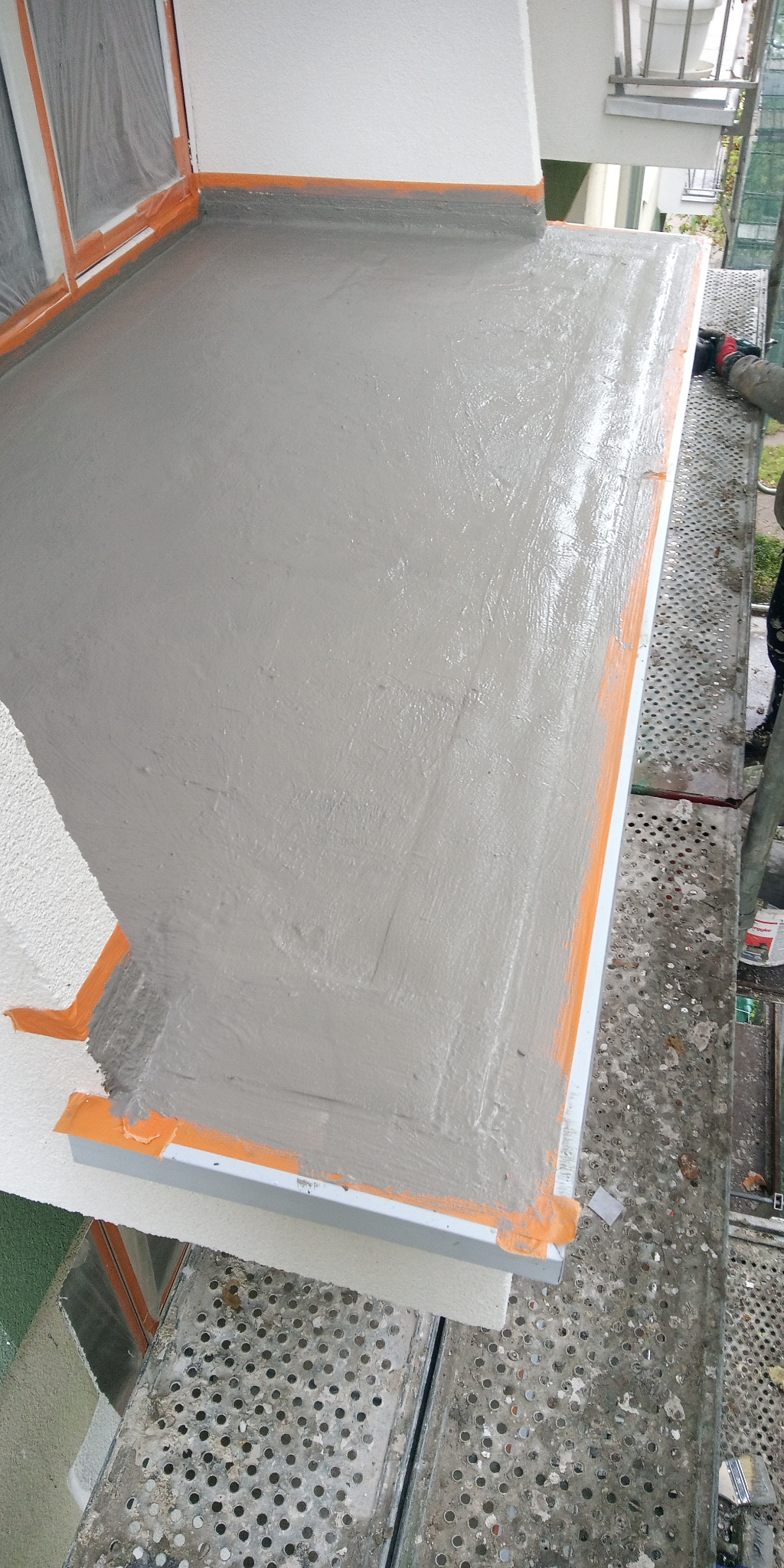 Szara, świeżo nałożona gładź na balkonie, zabezpieczona taśmą malarską, widok z góry, z fragmentem rusztowania i osoby w rękawicach w prawym górnym rogu.