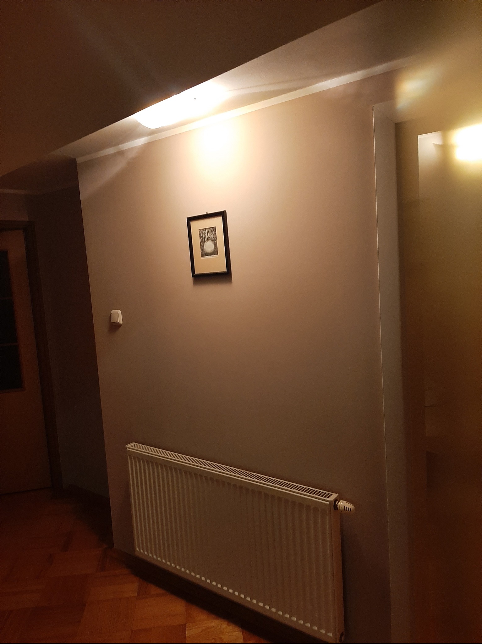 Beżowa ściana po malowaniu z grzejnikiem i obrazkiem w ramce. Widoczny fragment drewnianej podłogi i drzwi. Oświetlenie górne.