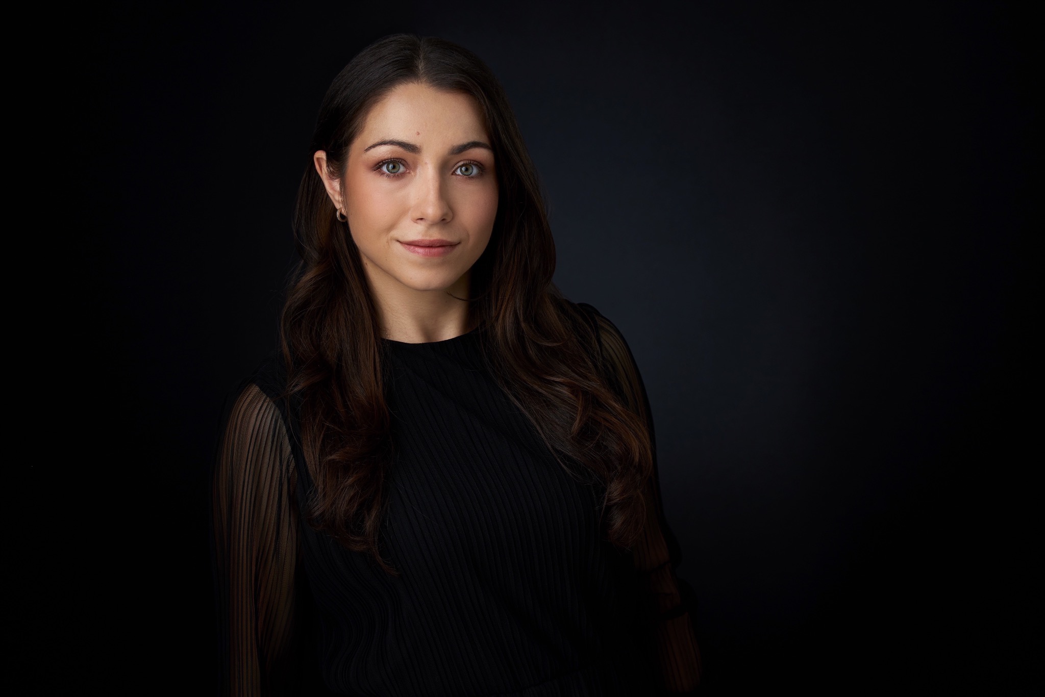 Portret młodej kobiety z długimi, brązowymi włosami i zielonymi oczami, ubrana w czarną bluzkę na ciemnym tle. Delikatny uśmiech, profesjonalny wygląd.