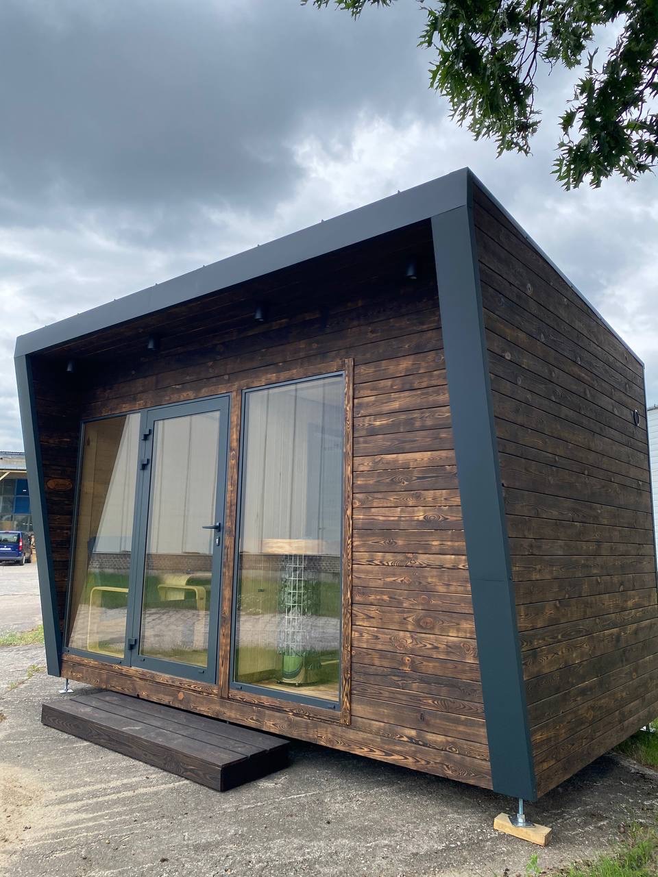 Nowoczesna, drewniana sauna zewnętrzna z dużymi przeszkleniami i ciemną, skośną konstrukcją dachu, ustawiona na betonowej powierzchni pod pochmurnym niebem.