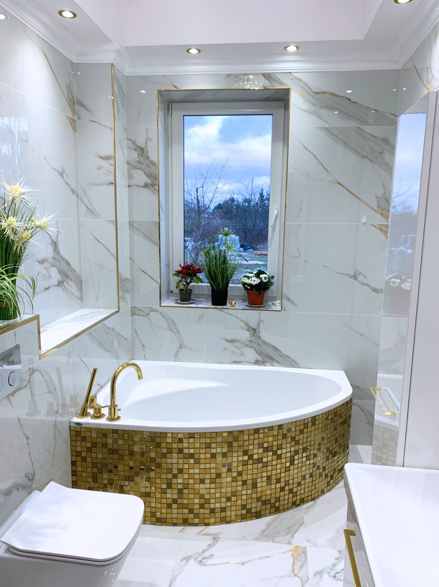 Elegancka łazienka z wanną obudowaną złotą mozaiką, marmurowe płytki z żyłkami złota, złota armatura, okno z roślinami. Luksusowe wykończenie.