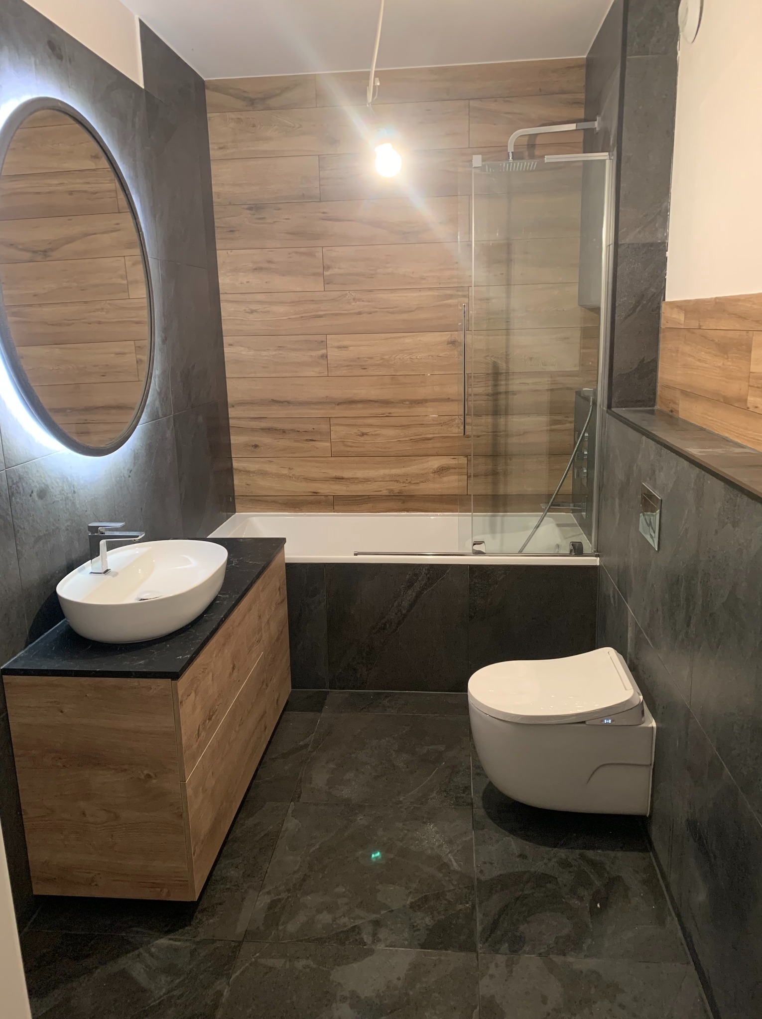 Nowoczesna łazienka z drewnopodobnymi płytkami, podświetlanym lustrem, umywalką nablatową i wiszącą toaletą. Ciemna, marmurkowa podłoga.