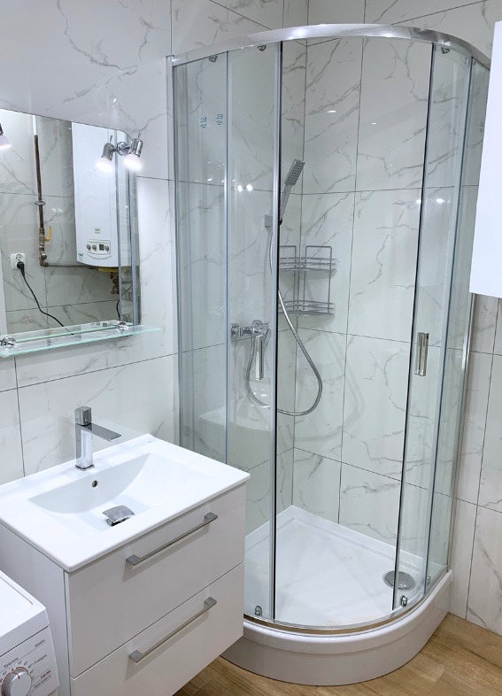 Jasna łazienka po remoncie: kabina prysznicowa narożna, umywalka z szafką, białe płytki imitujące marmur. Widoczny piec gazowy i pralka.