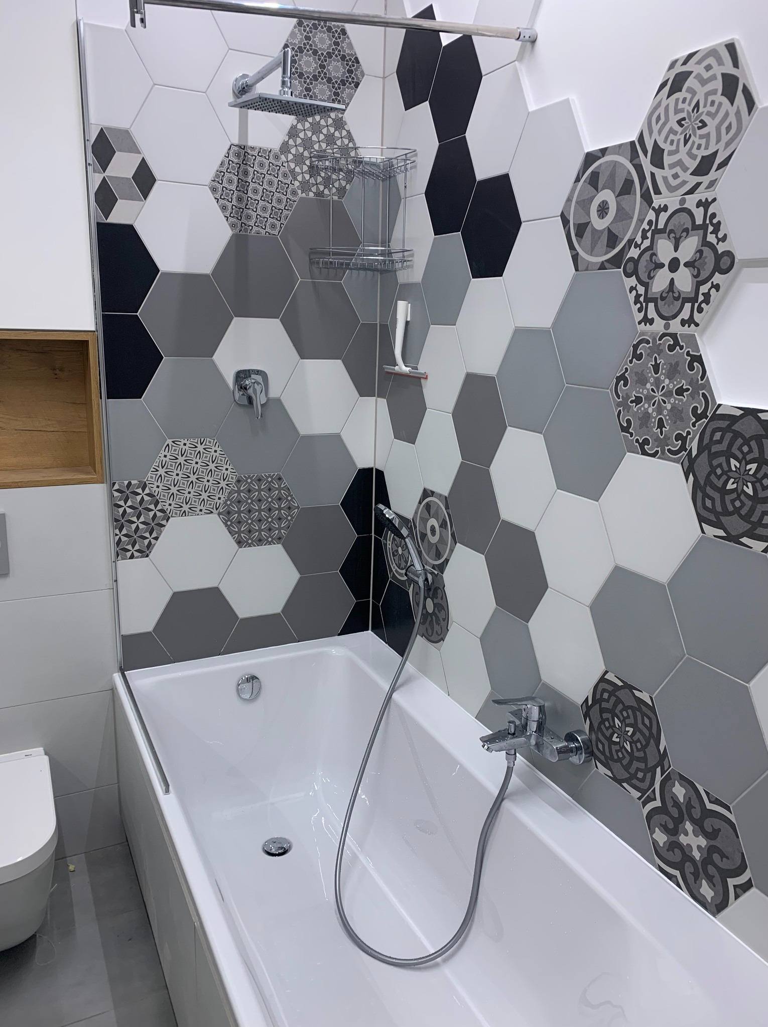 Nowoczesna łazienka z wanną, heksagonalne płytki w odcieniach szarości i bieli, akcenty wzorzyste. Chromowana armatura i minimalistyczna toaleta.