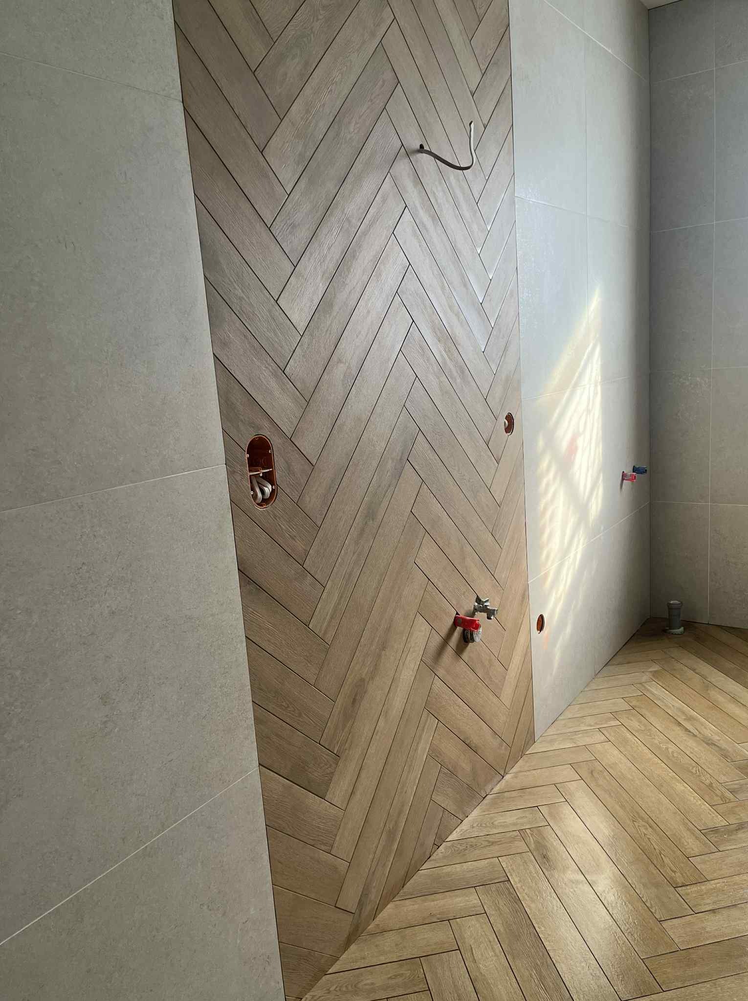 Układanie płytek drewnopodobnych w jodełkę na ścianie i podłodze łazienki. Widoczne przyłącza wodne i fragment ściany z szarych, matowych płytek.