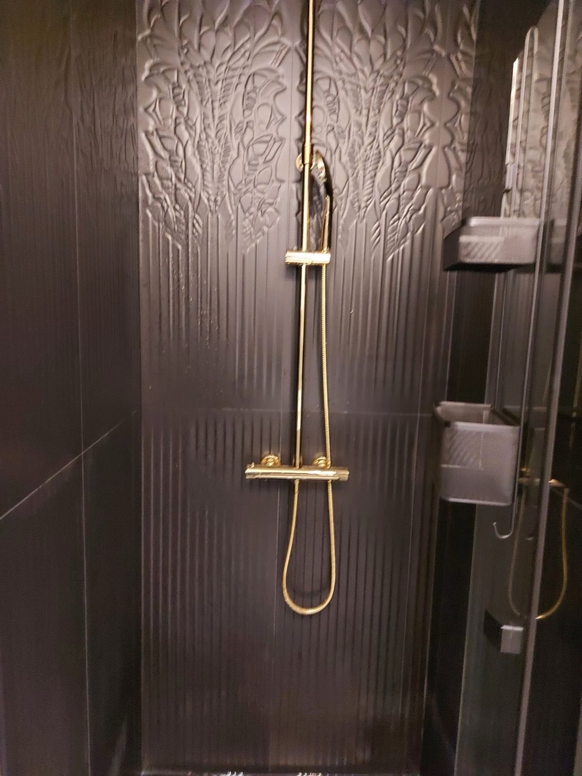Elegancka kabina prysznicowa z czarnymi płytkami o fakturze roślinnej i złotą armaturą. Nowoczesny design z półkami na kosmetyki.