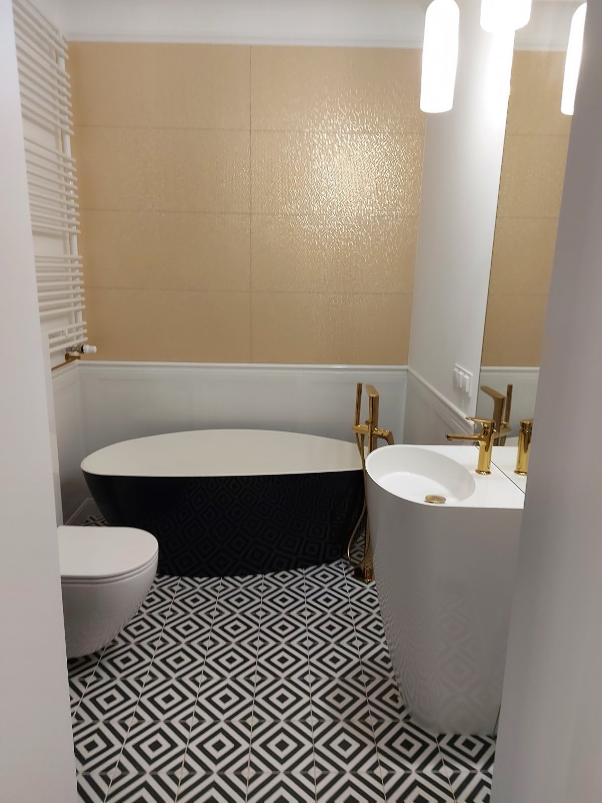 Elegancka łazienka z czarno-białą podłogą w geometryczny wzór, owalną wanną i umywalką na postumencie. Złota armatura i beżowe płytki dodają luksusowego charakteru.