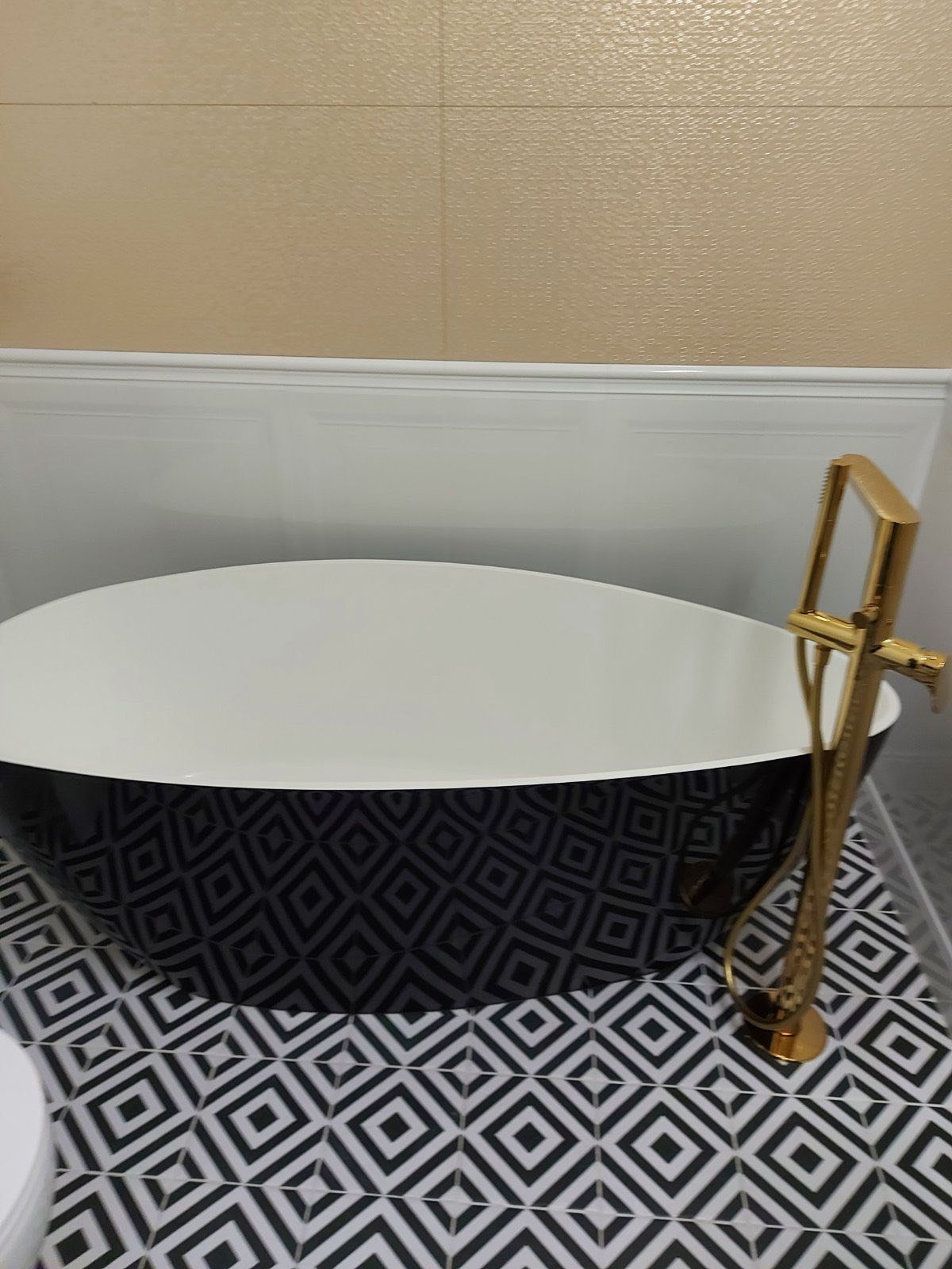 Elegancka, wolnostojąca wanna z czarno-białym wzorem na tle beżowych płytek i białej boazerii. Złota bateria dodaje luksusowego charakteru wykończonej łazience.