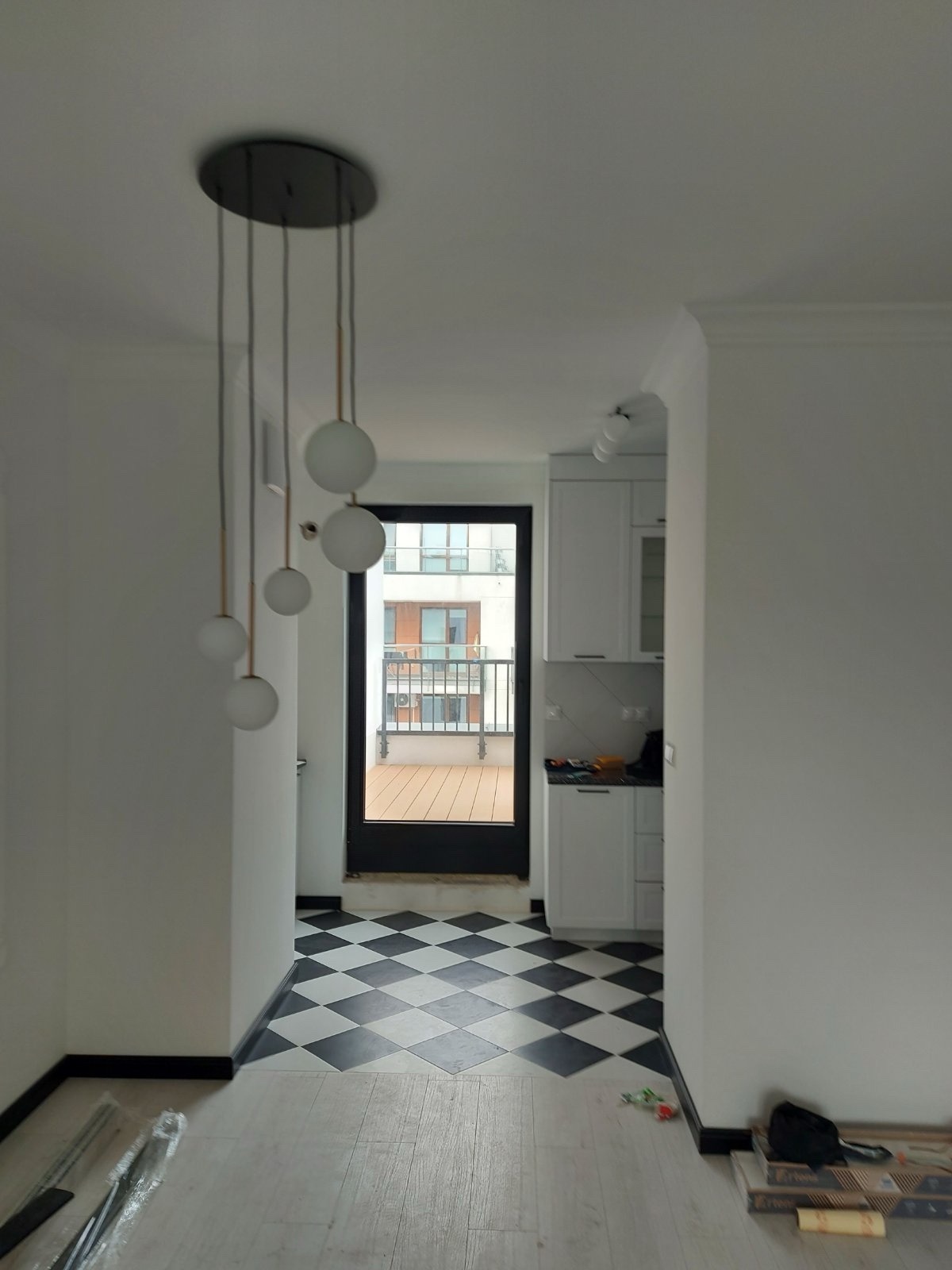 Wnętrze mieszkania po remoncie: białe ściany, czarno-biała podłoga w romby, nowoczesna lampa wisząca, widok na balkon i fragment białej zabudowy kuchennej.