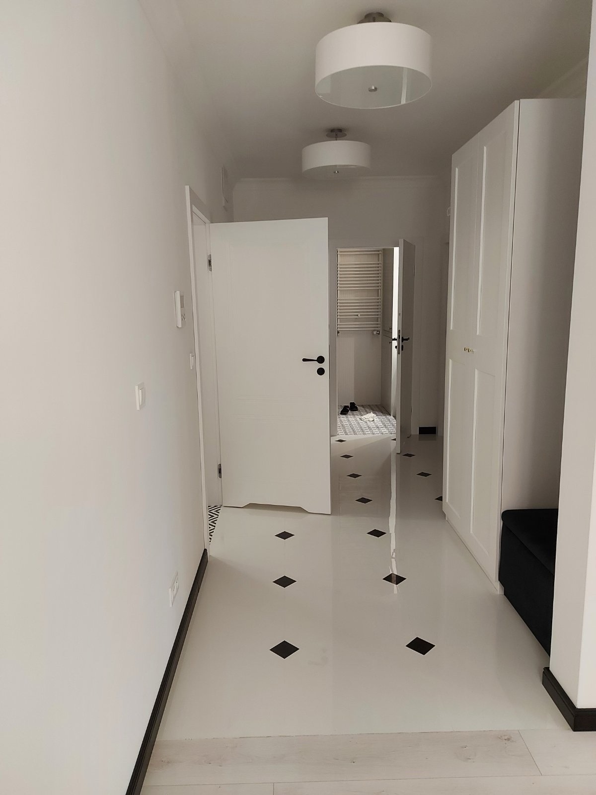 Biały korytarz z otwartymi drzwiami do łazienki, widoczna biała szafa i czarna pufa. Podłoga w jasnym odcieniu z czarnymi, romboidalnymi wzorami. Oświetlenie sufitowe.