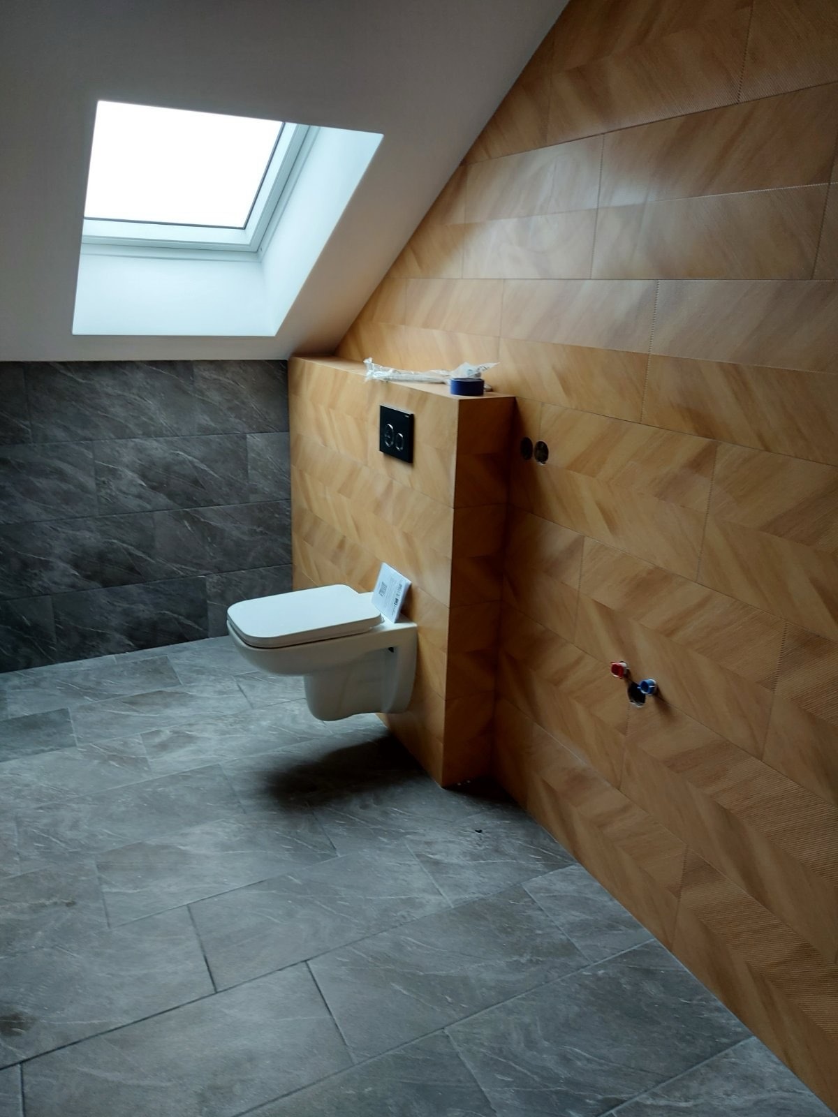 Nowoczesna łazienka na poddaszu: beżowe płytki w jodełkę, szare płytki imitujące kamień, podwieszana toaleta, okno dachowe. Minimalistyczny design.