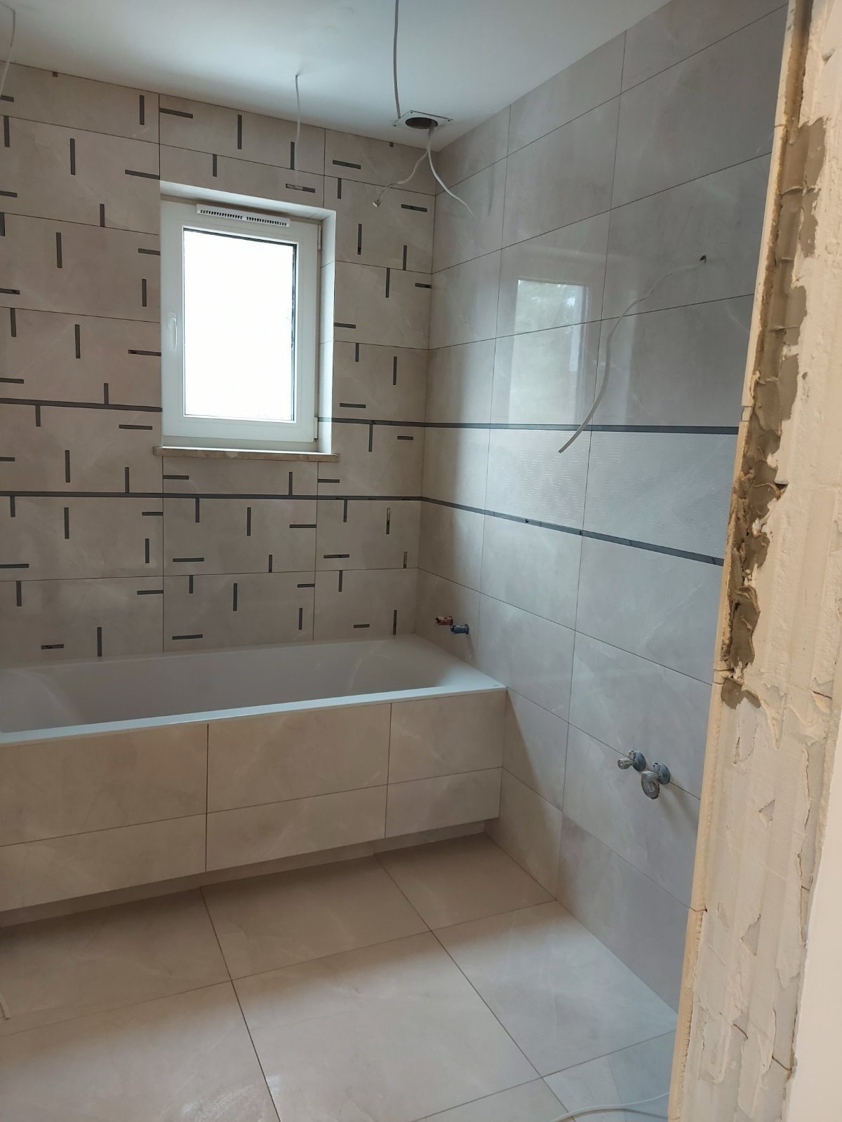 Wykończona łazienka z wanną w zabudowie, beżowe płytki z ciemnymi akcentami. Fragment ściany w surowym stanie, widoczne przewody elektryczne.