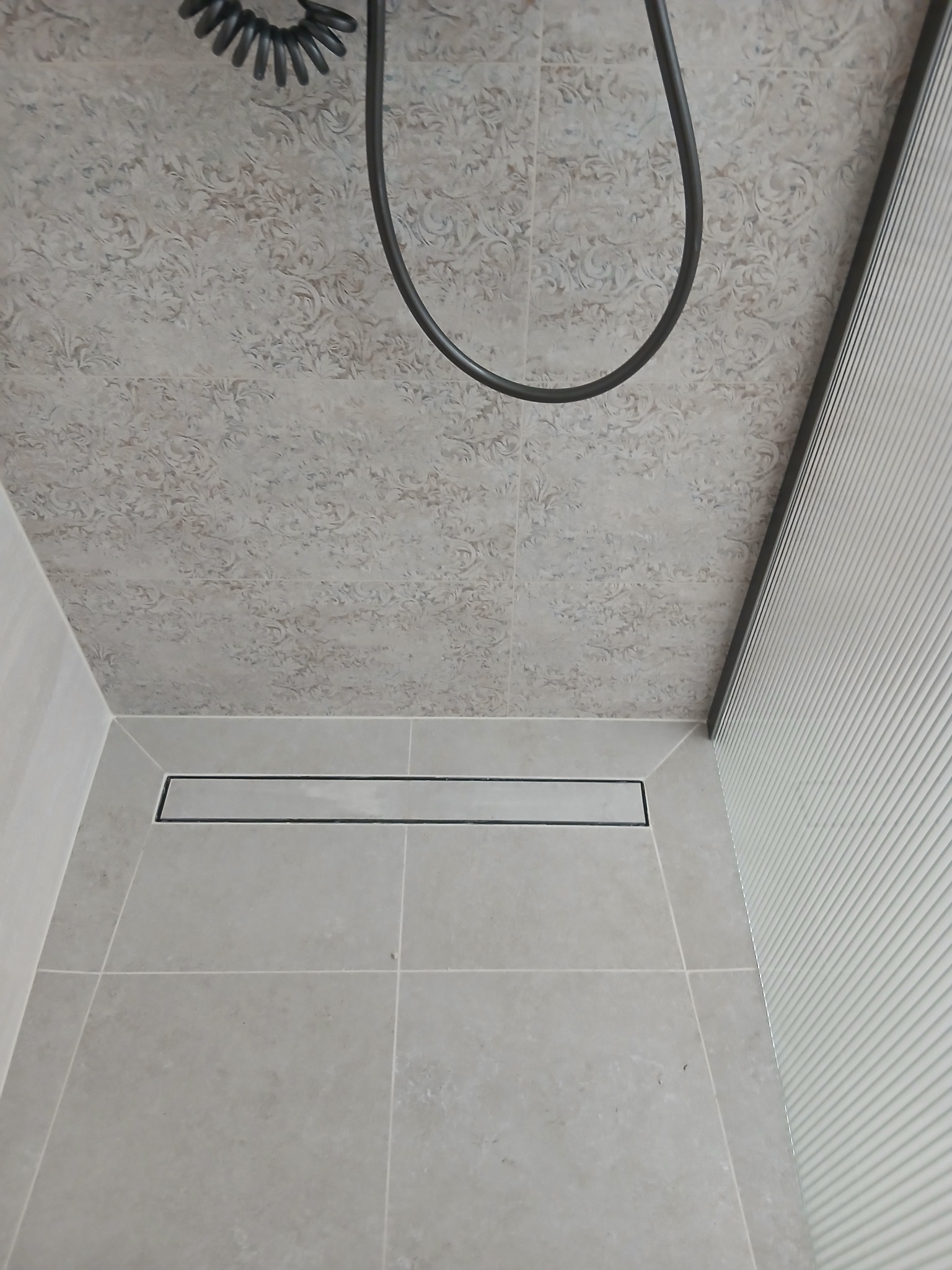 Wykończona kabina prysznicowa z odpływem liniowym, wyłożona jasnymi płytkami z subtelnym wzorem na ścianach i podłodze. Widoczny fragment słuchawki prysznicowej.