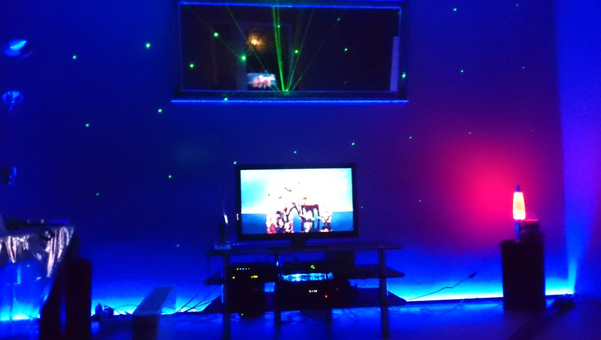 Pokój z efektem świetlnym: zielone lasery odbijają się w lustrze, ściany i meble oświetlone niebieskim światłem, lampka lawa dodaje czerwonego akcentu. Telewizor na środku.