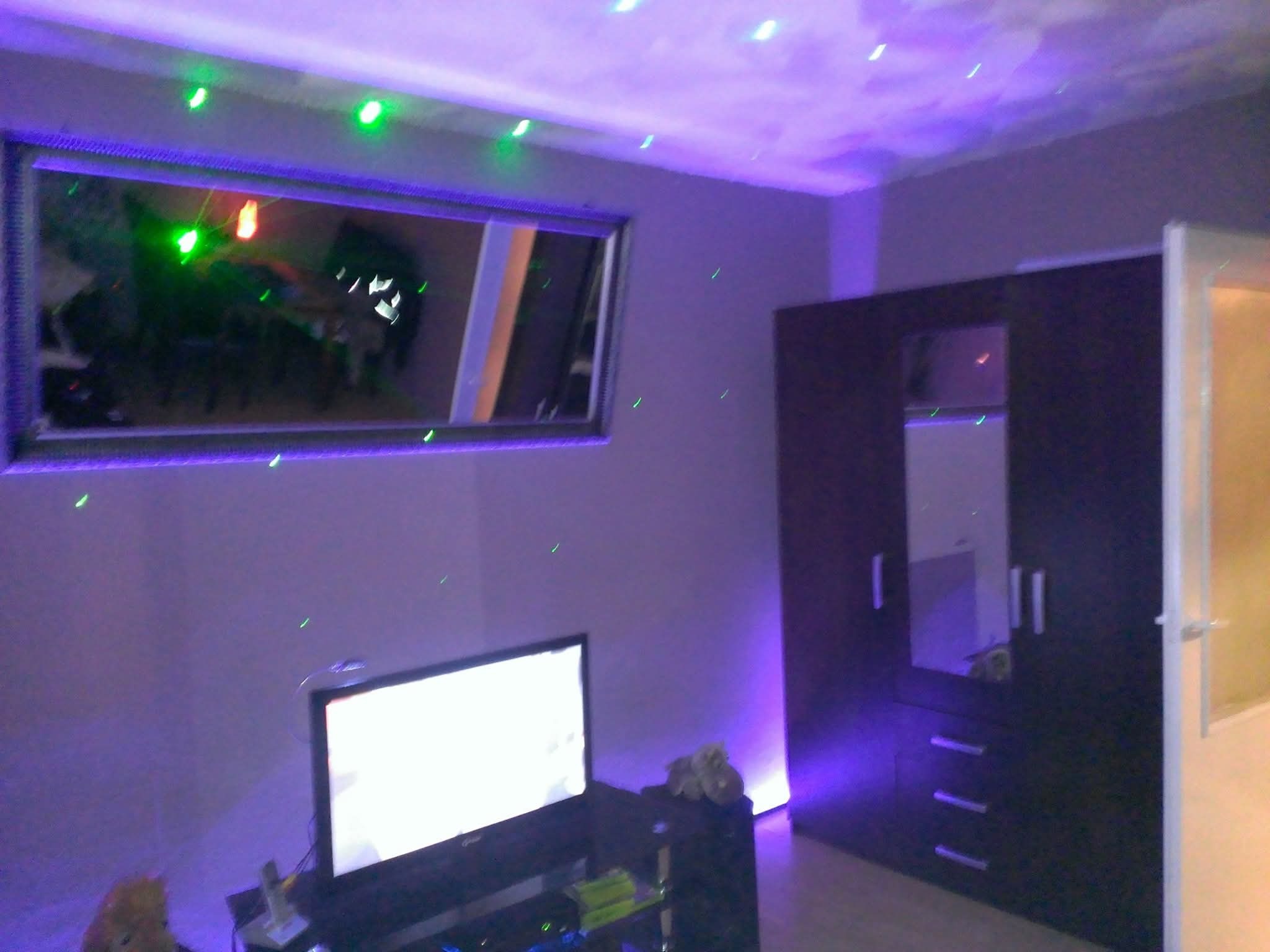 Pokój po remoncie: fioletowe oświetlenie LED, lustro w ramie, szafa z lustrem, telewizor na szafce. Zielone lasery na suficie dodają unikalnego charakteru.