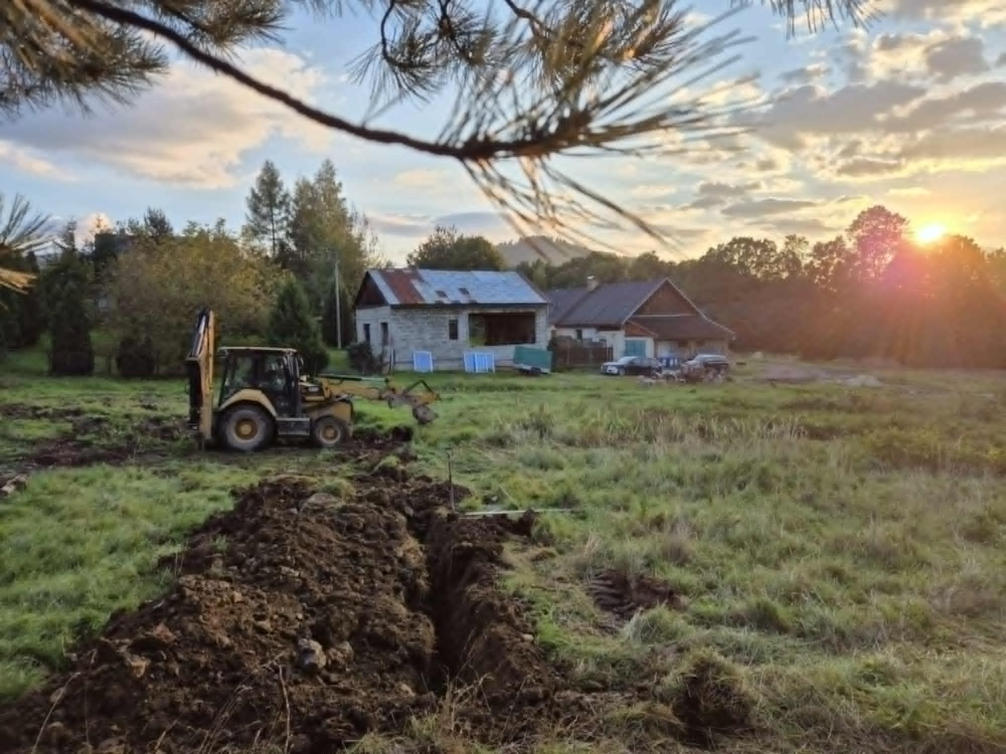 Żółta koparko-ładowarka w trakcie prac ziemnych na zalesionym terenie, wykopany rów, w tle budynki mieszkalne i zachodzące słońce.