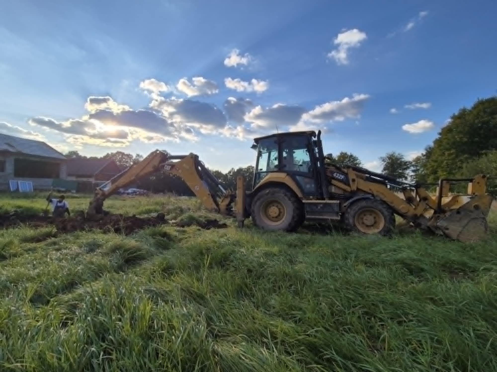 Żółta koparko-ładowarka CAT 432F w trakcie prac ziemnych na zarośniętym terenie, w tle widoczny budynek i zachodzące słońce.