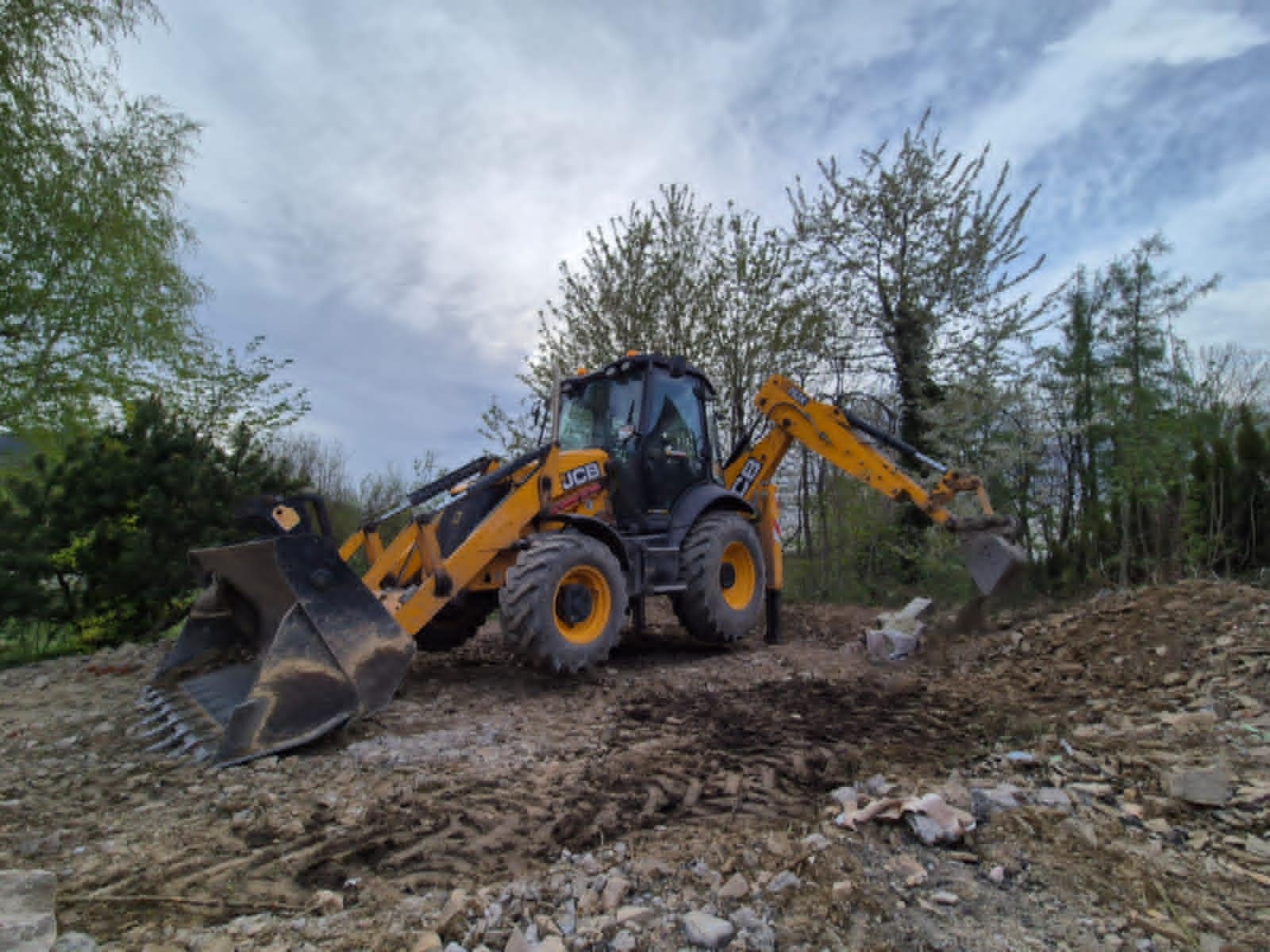 Żółta koparko-ładowarka JCB w trakcie pracy na placu budowy, z widocznym wykopem i gruzem. W tle drzewa i pochmurne niebo.