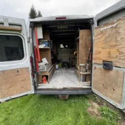 Otwarty tył furgonetki z narzędziami i sprzętem na tle trawnika, sugerujący gotowość do pracy. Widoczne p&oacute;łki i uporządkowane narzędzia.