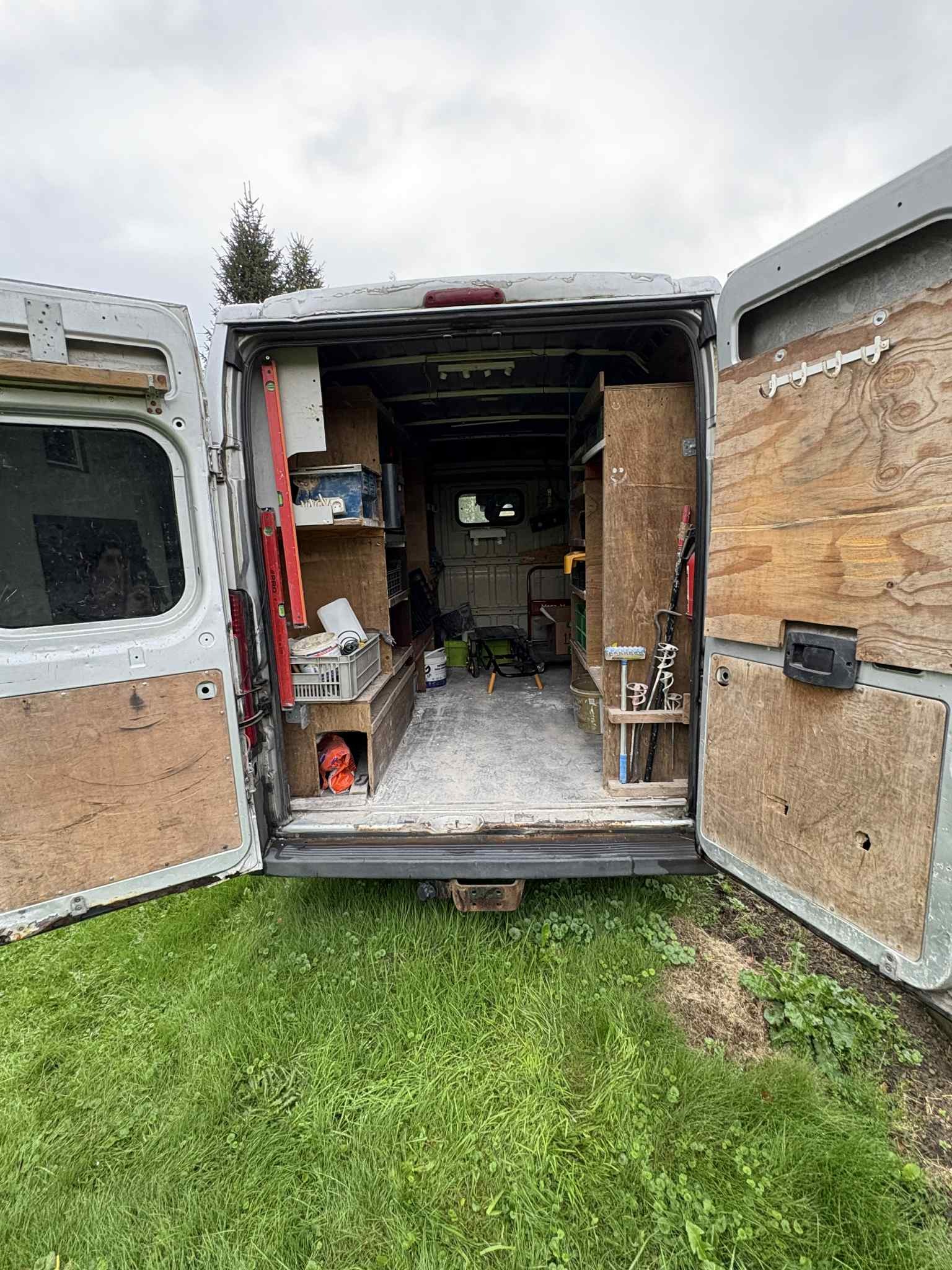Otwarty tył furgonetki z narzędziami i sprzętem na tle trawnika, sugerujący gotowość do pracy. Widoczne półki i uporządkowane narzędzia.