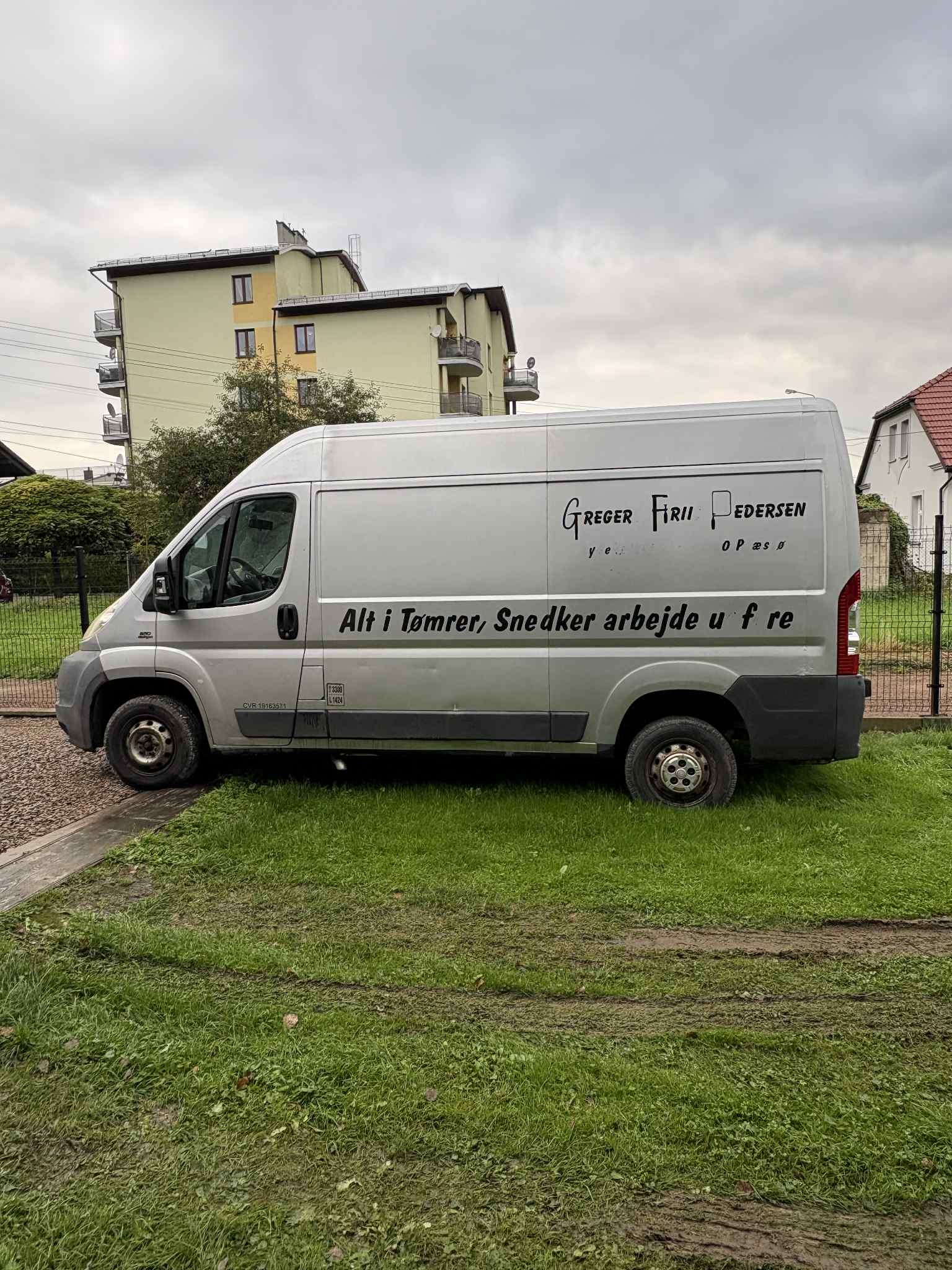 Srebrny bus firmy remontowej Greger Firii Pedersen z napisem 'Alt i Tømrer, Snedker arbejde u f re' na tle budynku mieszkalnego i trawnika.