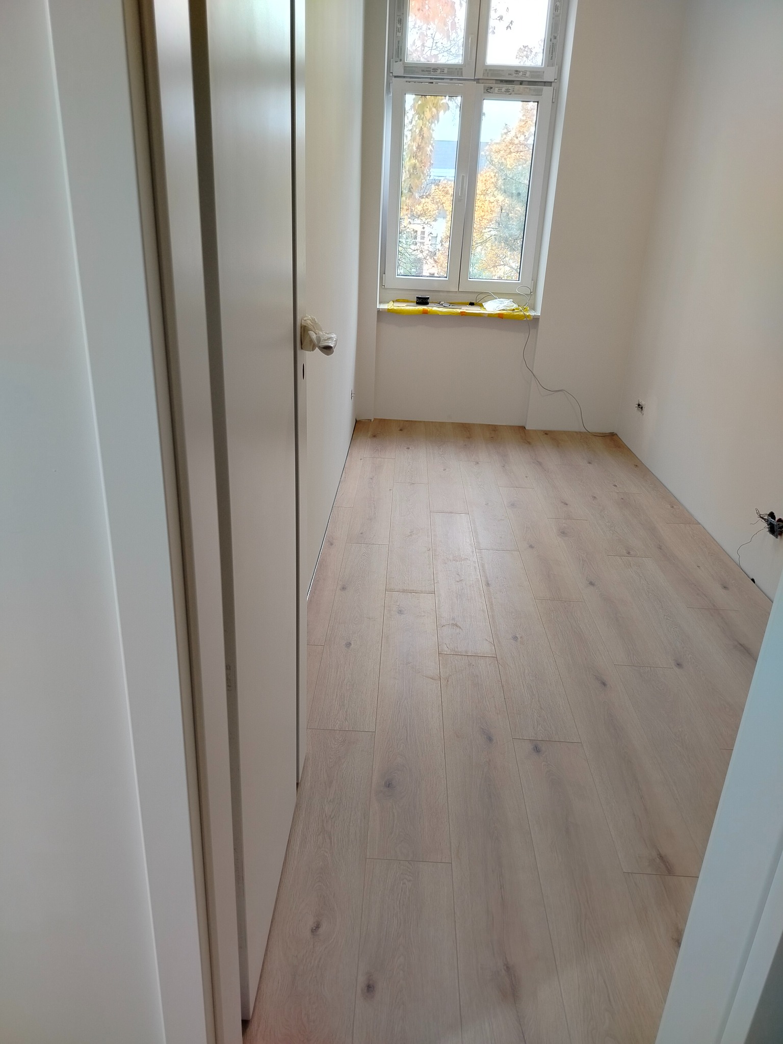 Jasne panele podłogowe w pokoju po remoncie, widok zza otwartych, białych drzwi. Okno z widokiem na drzewa w tle. Gniazdka elektryczne na ścianie.