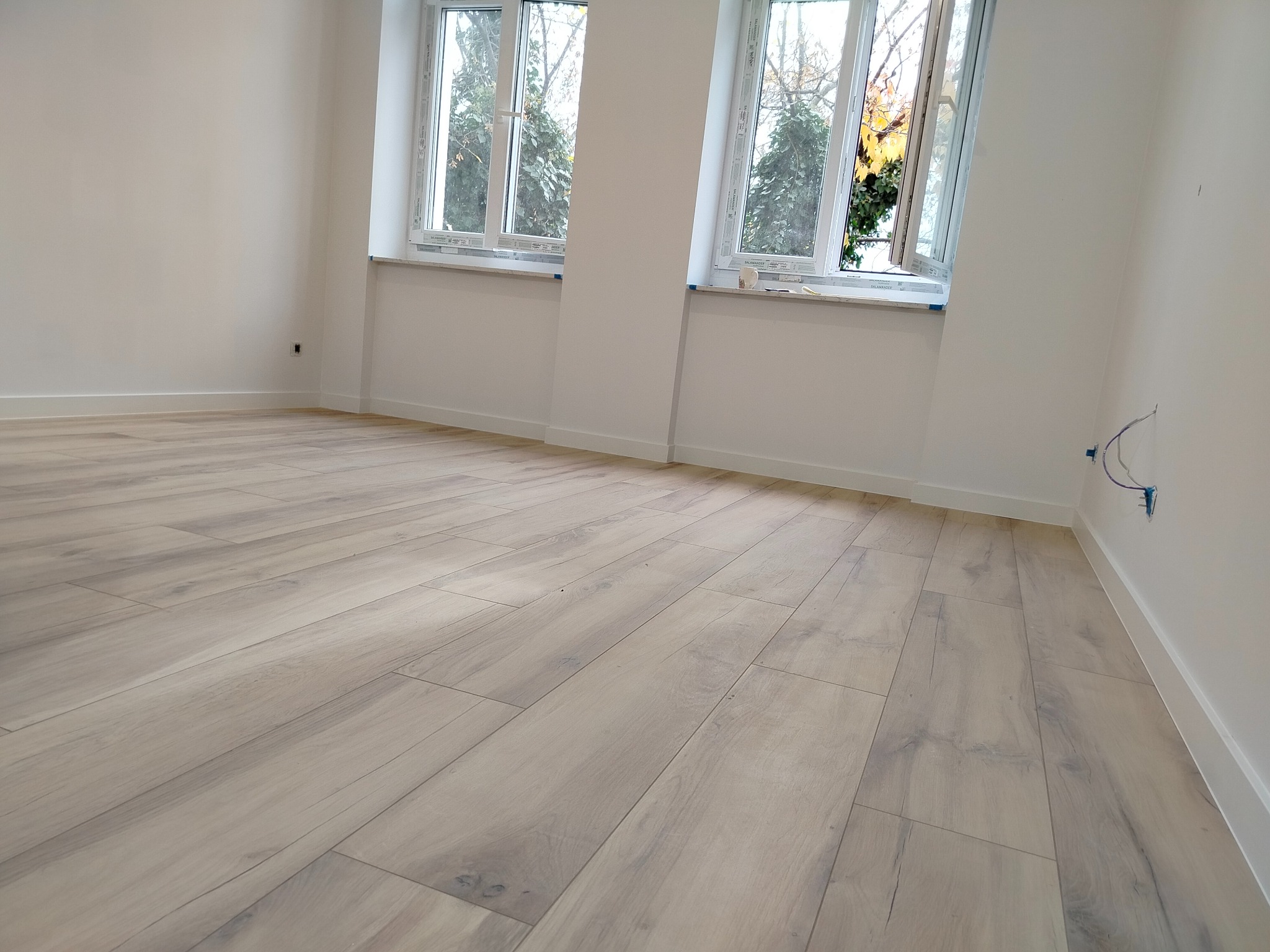 Jasne panele podłogowe ułożone w pustym, odnowionym pokoju z dwoma oknami. Świeżo pomalowane ściany i białe listwy przypodłogowe. Widoczne gniazdka elektryczne.