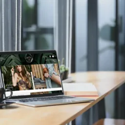 Laptop prezentuje stronę sklepu odzieżowego z motywami roślinnymi, obok filiżanka kawy i książka na drewnianym stole, w tle okno z widokiem na zieleń.