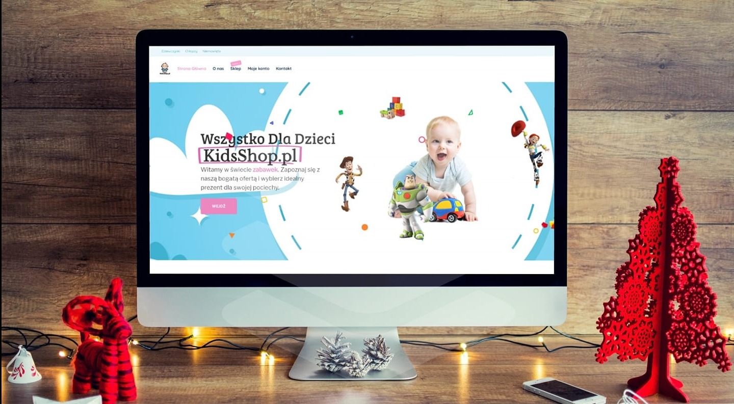 Ekran komputera wyświetla stronę sklepu internetowego KidsShop.pl z zabawkami dla dzieci. W tle drewniana ściana i świąteczne dekoracje, tworzące przytulny nastrój.