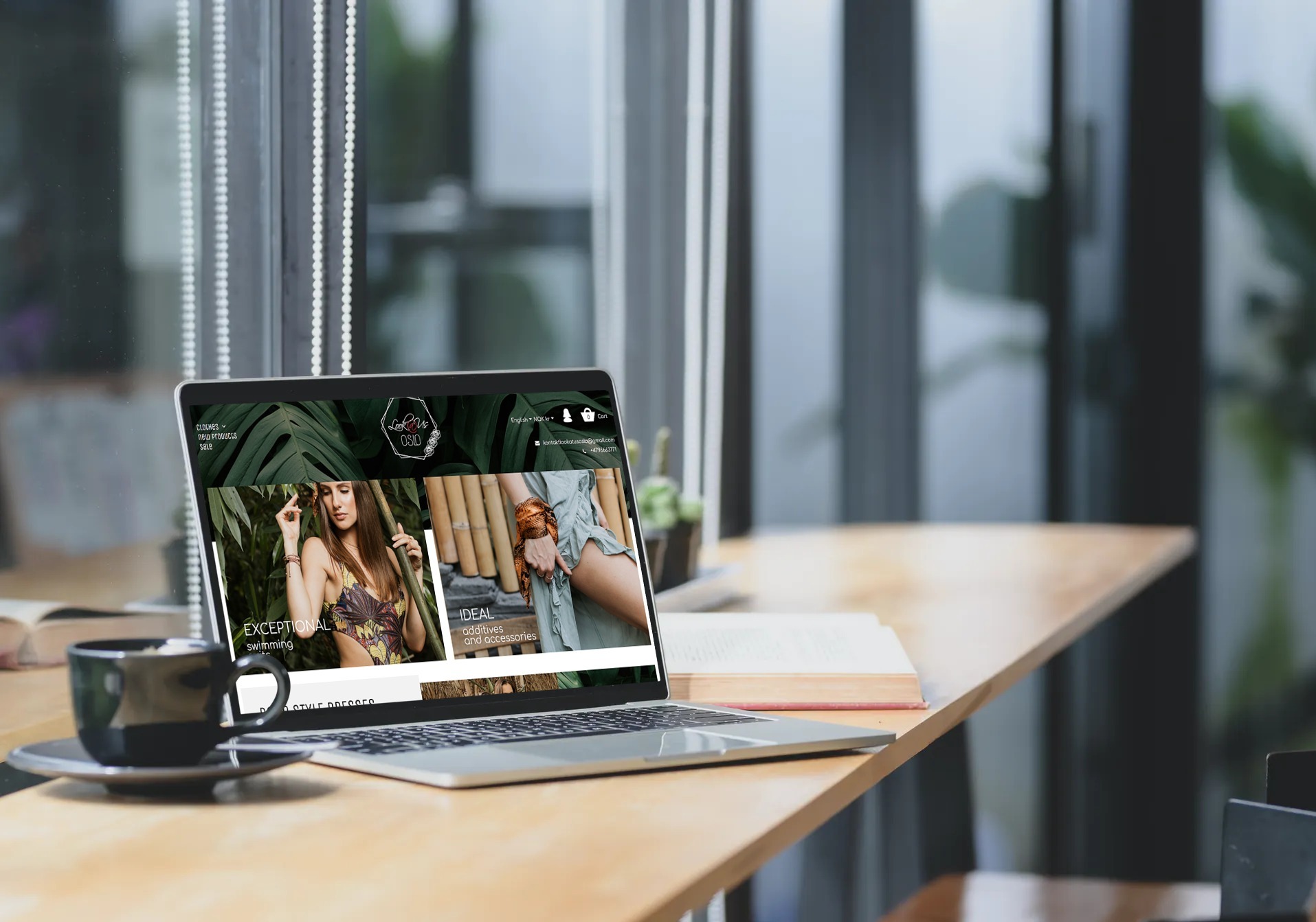 Laptop prezentuje stronę sklepu odzieżowego z motywami roślinnymi, obok filiżanka kawy i książka na drewnianym stole, w tle okno z widokiem na zieleń.
