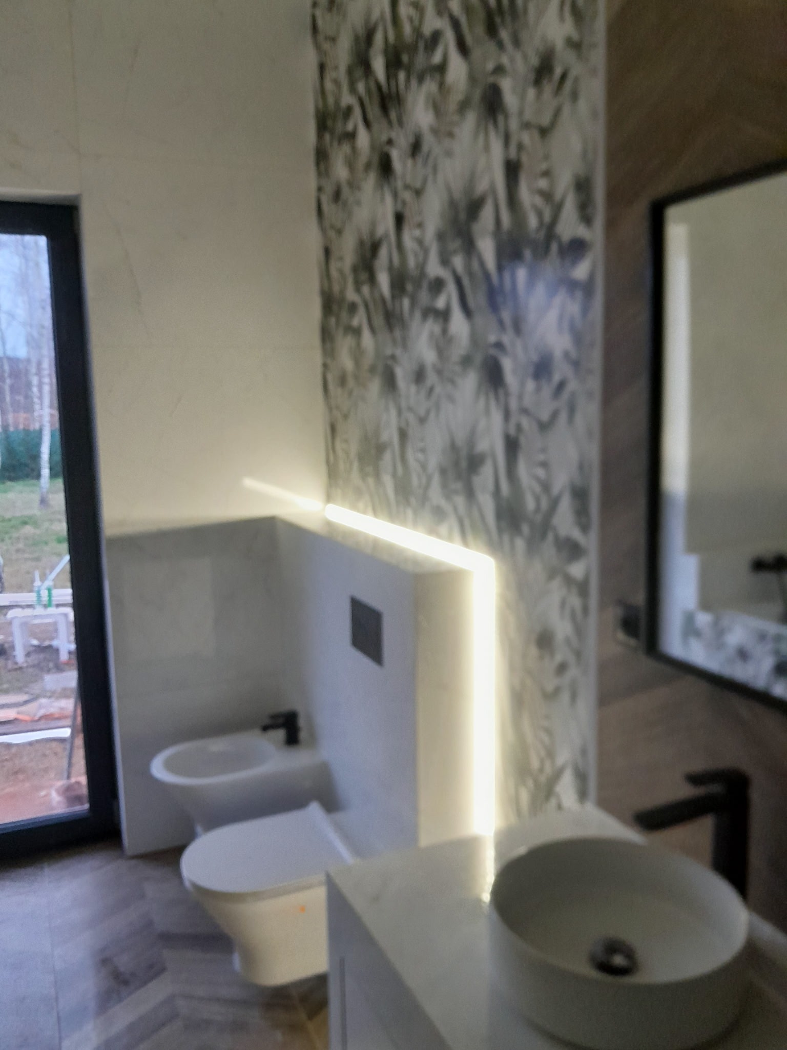 Nowoczesna łazienka z podświetlanym panelem, umywalką nablatową i wzorzystą tapetą. Bidet i toaleta w minimalistycznym stylu. Widok z zewnątrz przez okno.