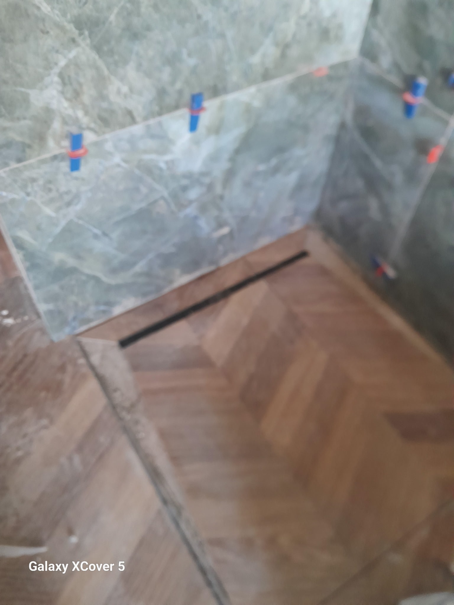 Fragment łazienki w trakcie wykańczania: ułożone płytki marmurowe na ścianach z niebieskimi krzyżykami dystansowymi, drewnopodobna podłoga w jodełkę i odpływ liniowy.