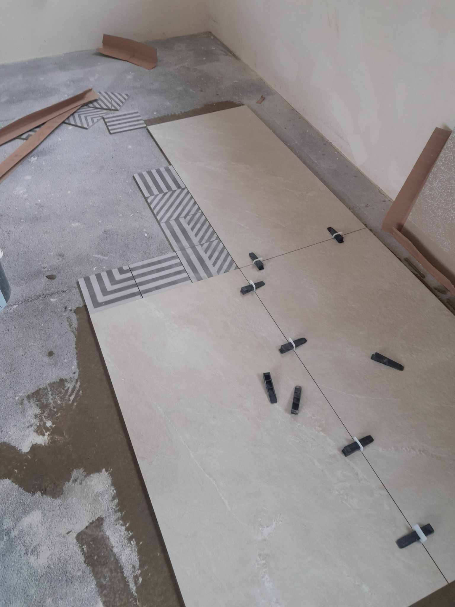 Układanie beżowych płytek z wzorem, z użyciem krzyżyków dystansowych. Częściowo ułożone płytki z geometrycznym wzorem. Widoczny fragment podłogi w trakcie prac.