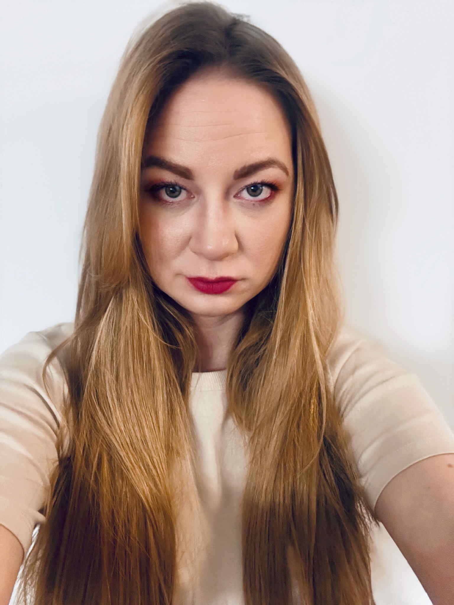 Portret kobiety z długimi, blond włosami i wyrazistym makijażem oczu oraz ust. Ujęcie z bliska, na neutralnym tle. Widoczny fragment ramienia w jasnej bluzce.