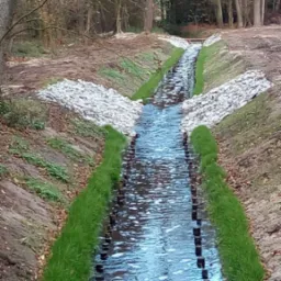 Uregulowany rów melioracyjny z drewnianymi palikami i obsianą trawą skarpą, wzmocniony kamieniami, w tle las