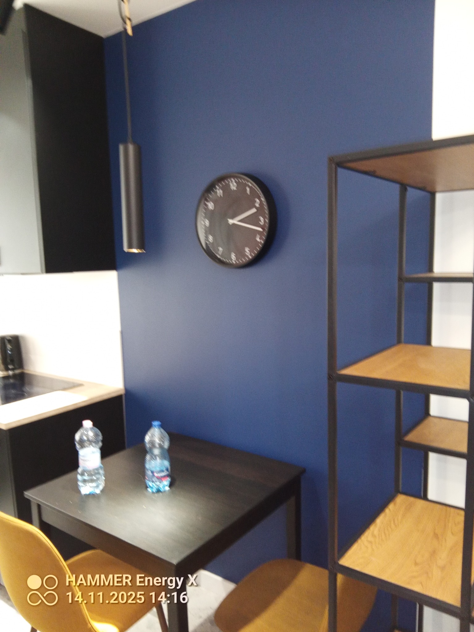 Nowoczesna kuchnia z granatową ścianą, zegarem i minimalistyczną lampą wiszącą. Na stole dwie butelki wody. W tle czarne szafki i metalowy regał.