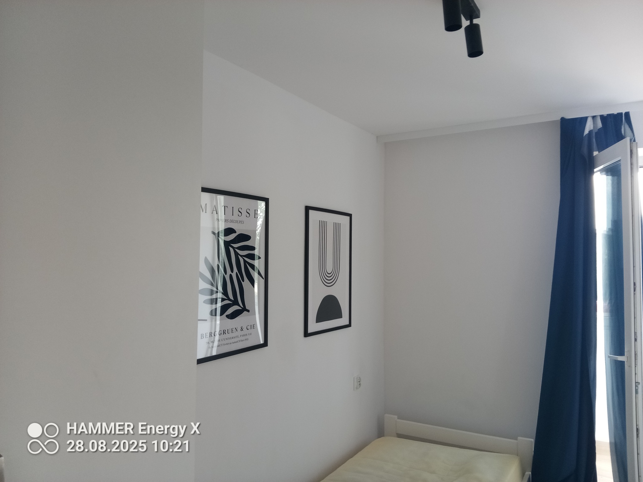 Minimalistyczna sypialnia z dwoma czarno-białymi plakatami w ramkach na białej ścianie, lampą sufitową i zasłoniętym oknem balkonowym. Jasne wnętrze.