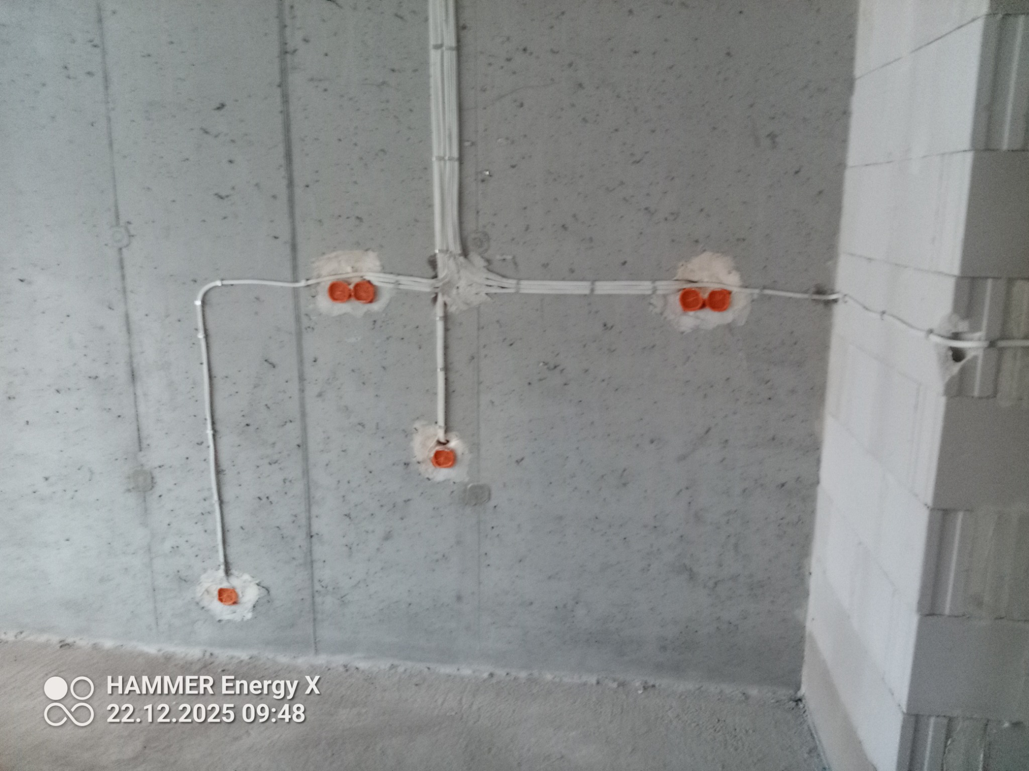 Szare ściany w surowym stanie z widocznymi, zamontowanymi przewodami elektrycznymi i pomarańczowymi puszkami pod gniazdka. Widoczne łączenia kabli.