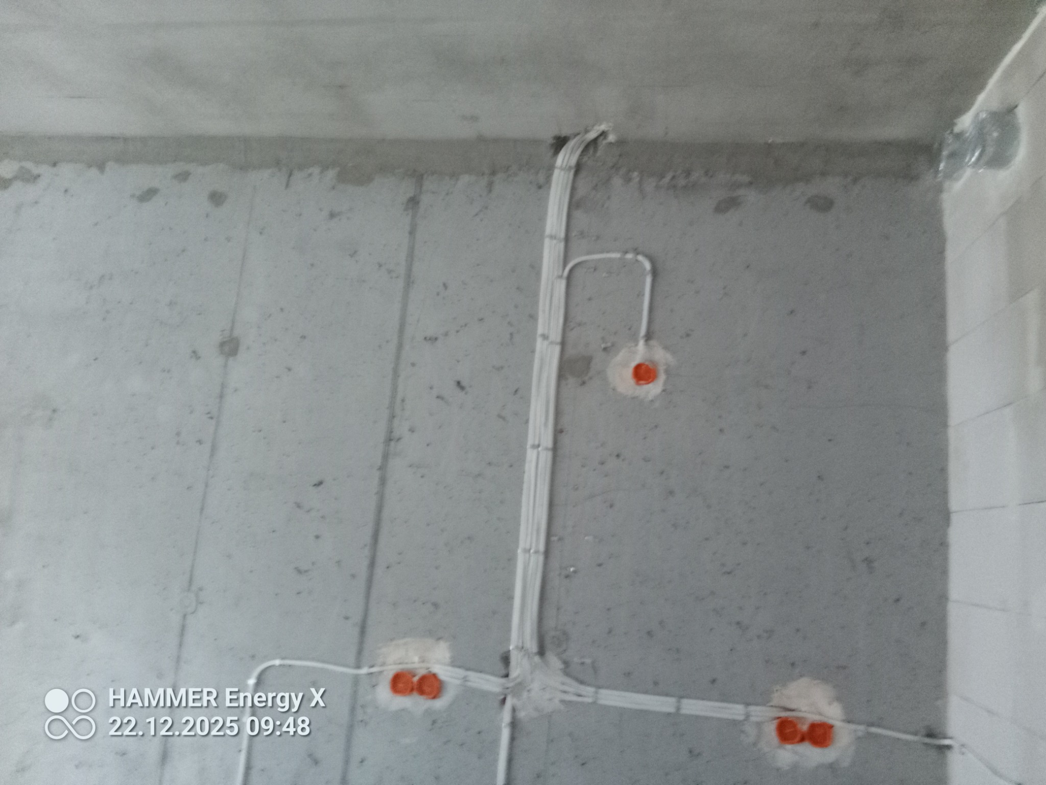Surowa ściana z widoczną, częściowo zamontowaną instalacją elektryczną w białych rurkach i pomarańczowych puszkach. Widok z dołu, sufit betonowy.