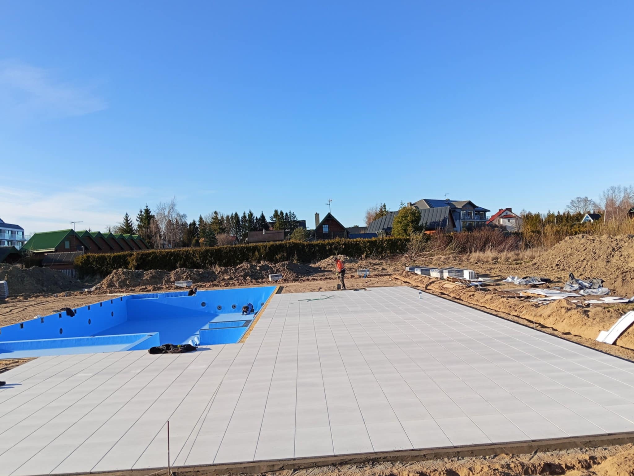 Błękitny basen w trakcie budowy, otoczony jasnoszarą kostką brukową. W tle domy i błękitne niebo. Prace brukarskie w toku, widoczny pracownik przy basenie.