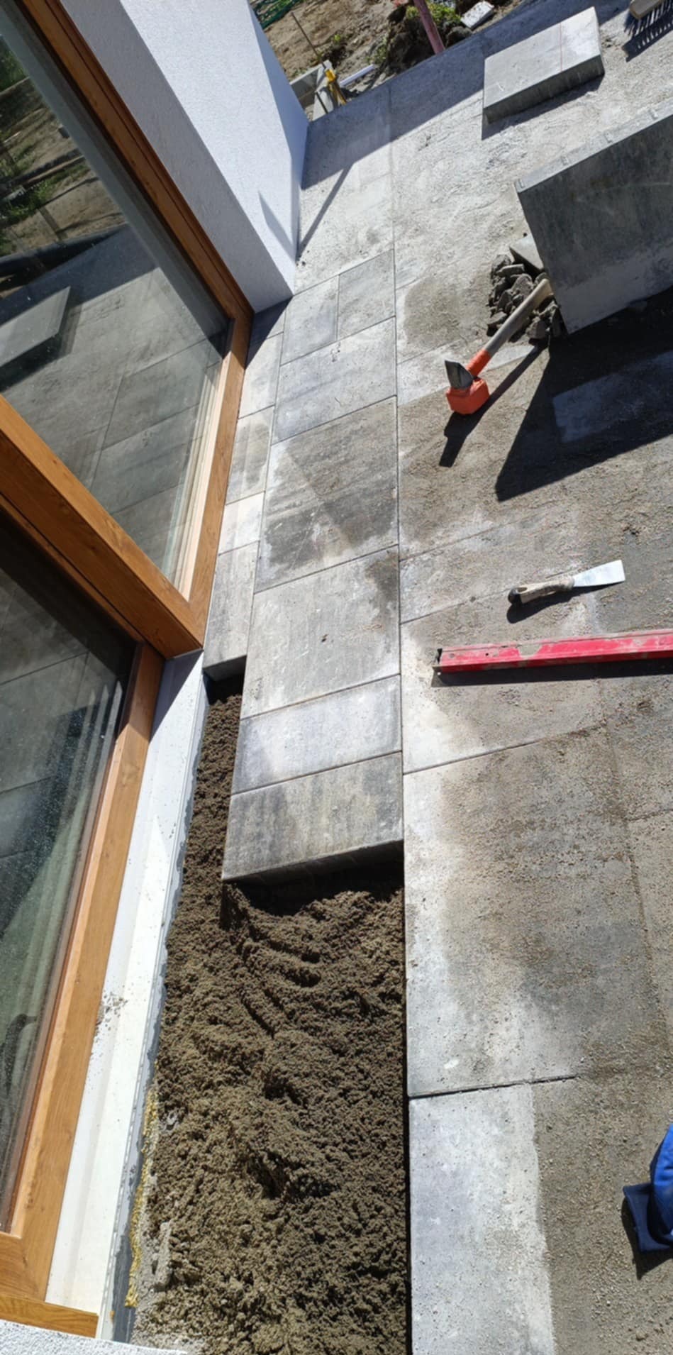 Układanie kostki brukowej przy oknie domu. Widoczne narzędzia: młotek, poziomica. Częściowo ułożona kostka i piasek. Kątowe ujęcie z góry.
