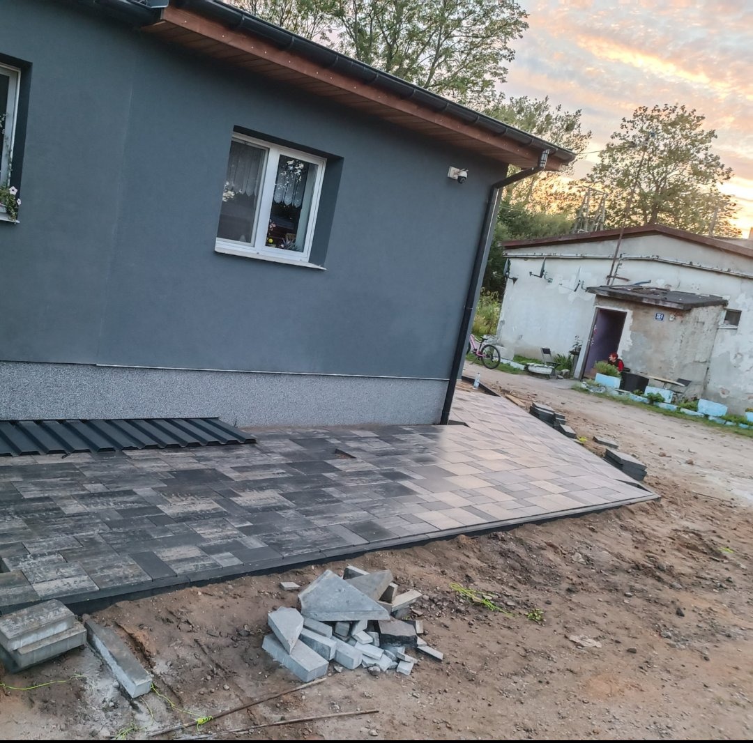 Ułożona kostka brukowa w odcieniach szarości przy szarym domu z białymi oknami, z elementami luźnej kostki na pierwszym planie i budynkiem gospodarczym w tle.