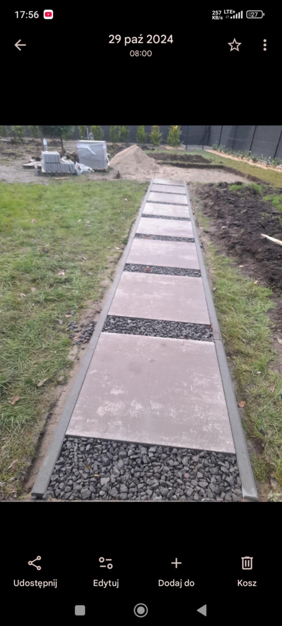 Ścieżka z płyt betonowych i grysu, obrzeża betonowe, trawnik po bokach. W tle hałda piasku i kostka brukowa. Ujęcie z perspektywy pieszego.