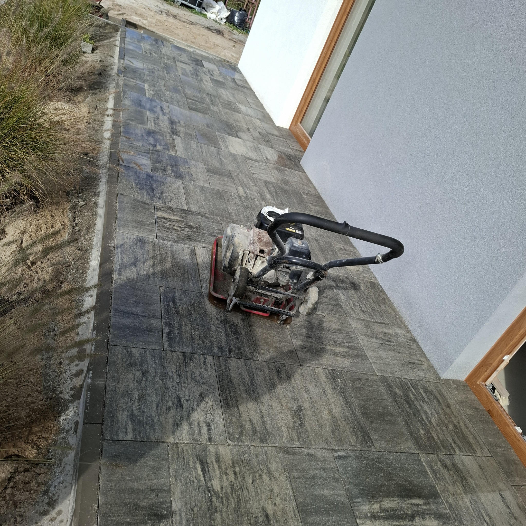 Układanie kostki brukowej w Białogardzie. Wibro ubijak na ciemnoszarej kostce brukowej. Widoczny fragment budynku i trawa.