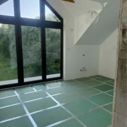 Pokój na poddaszu z dużym oknem i zieloną podłogą przygotowaną do układania parkietu. Widoczny surowy betonowy element konstrukcyjny.